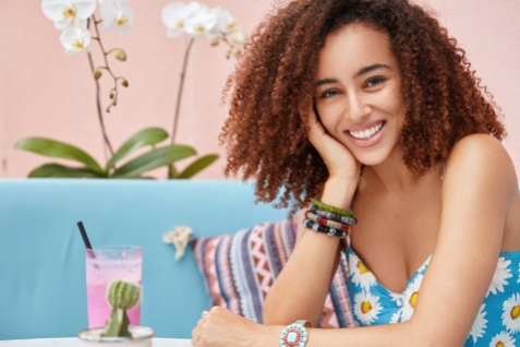 Positive dark skinned curly female with broad smile enjoys good recreation in cafeteria, drinks cocktail while sits on comfortable couch against pink wall with orchid in background, has rest