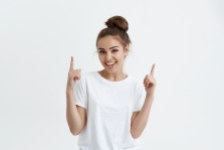 Positive european woman pointing up with both index fingers while smiling cheerfully at camera, standing over white background. Modern woman brags about her new ceiling that was made by famous painter.