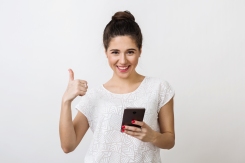 stylish young woman with attractive face holding smartphone and showing thumb up, positive gesture, smiling, good mood, using mobile device, isolated on white background